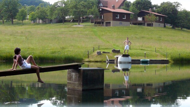 A serene lakeside scene with a person sitting on a dock, another standing on a platform, and a building in the background. - 2 Siefertshof Comfortable Holiday Residence (Mossautal)