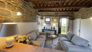 A cozy living room with stone walls, wooden beams, two sofas, a table with a lamp, and a view through a glass door.
