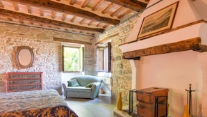 A rustic bedroom with exposed wooden beams, stone walls, a bed with a patterned bedspread, a checkered sofa, a dresser, and a fireplace.