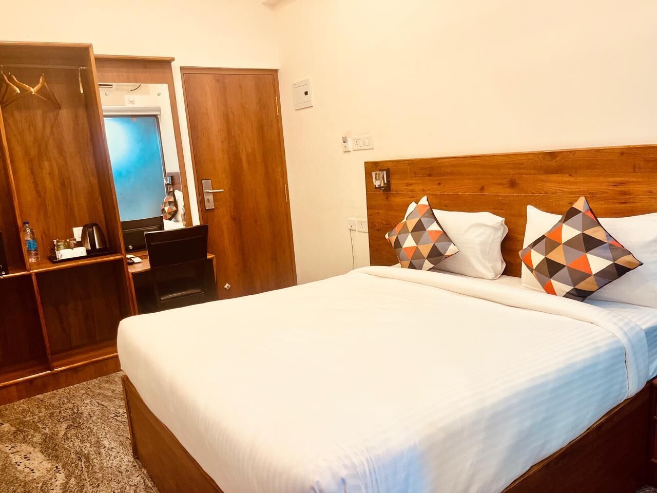 A hotel room with a wooden headboard, a bed with white linens, two patterned pillows, a desk with a laptop, and a wardrobe with a mirror.