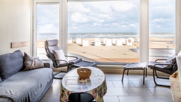 Ett rum med ett stort fönster med utsikt över en strand, en soffa, två fåtöljer och ett litet bord med en korg.