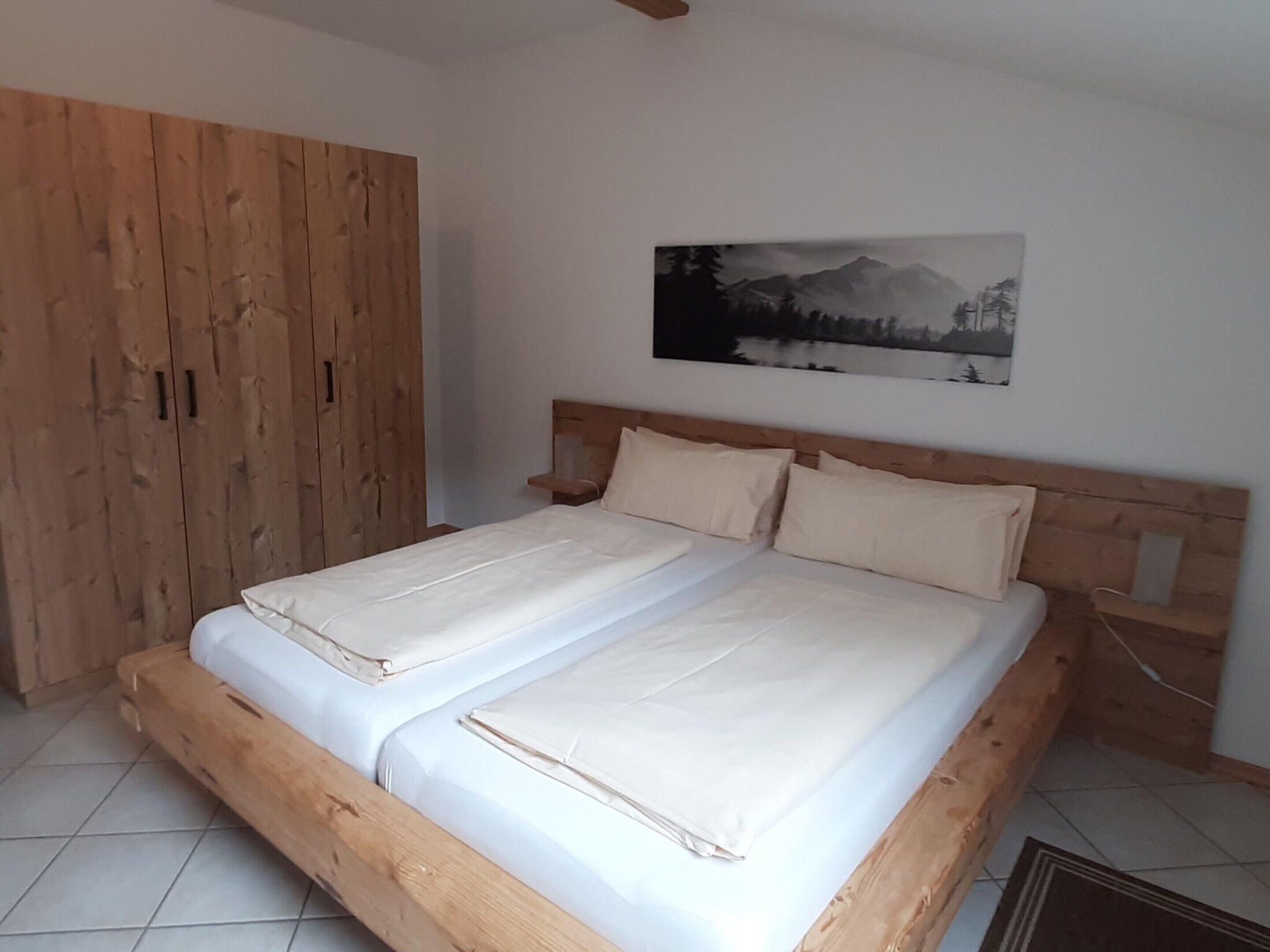 A bedroom with a wooden bed frame, two pillows, and a wall-mounted painting of a mountain landscape.