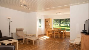 A spacious living room with a wooden floor, a dining area with a wooden table and chairs, a sofa, and a TV on a wooden cabinet.
