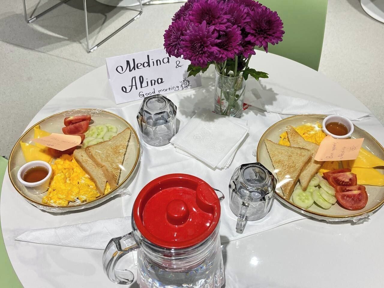 Una mesa de desayuno con dos platos de comida, una taza roja y un jarrón con flores moradas.