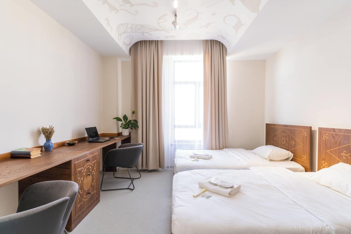 Habitación de hotel con un escritorio de madera, dos camas, una silla y un ventanal con cortinas.