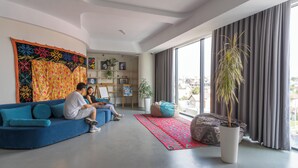 Un salon moderne avec un canapé bleu, une grande tapisserie colorée, une plante en pot et une vue sur la ville à travers de grandes fenêtres.