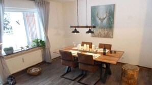 A dining area with a wooden table, chairs, and hanging pendant lights. There is a radiator, a window with curtains, and a painting on the wall. - Nice Apartment in Hahnenklee (Goslar)