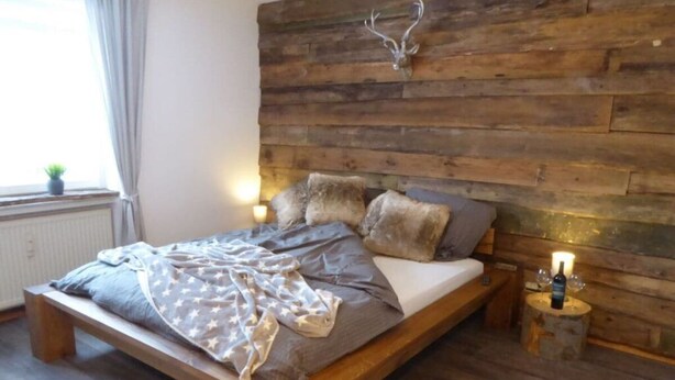 A bed with a wooden headboard, a bench, and a bedside table with a bottle and glasses. - Nice Apartment in Hahnenklee (Goslar)