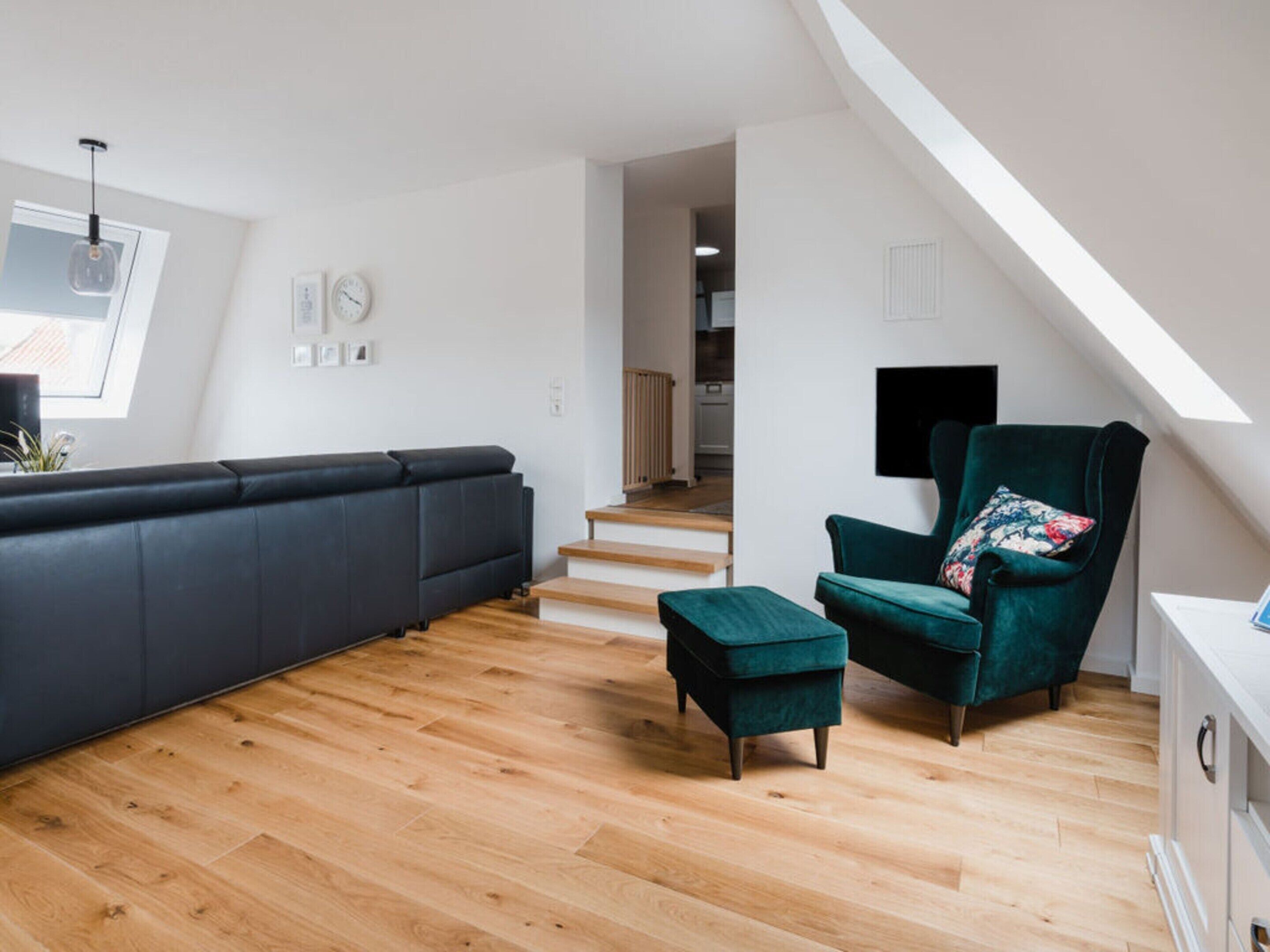 A modern living room with a dark grey sectional sofa, a teal armchair with a patterned cushion, and a matching ottoman. The room features a wooden floor, a staircase leading to an upper level, and a skylight.