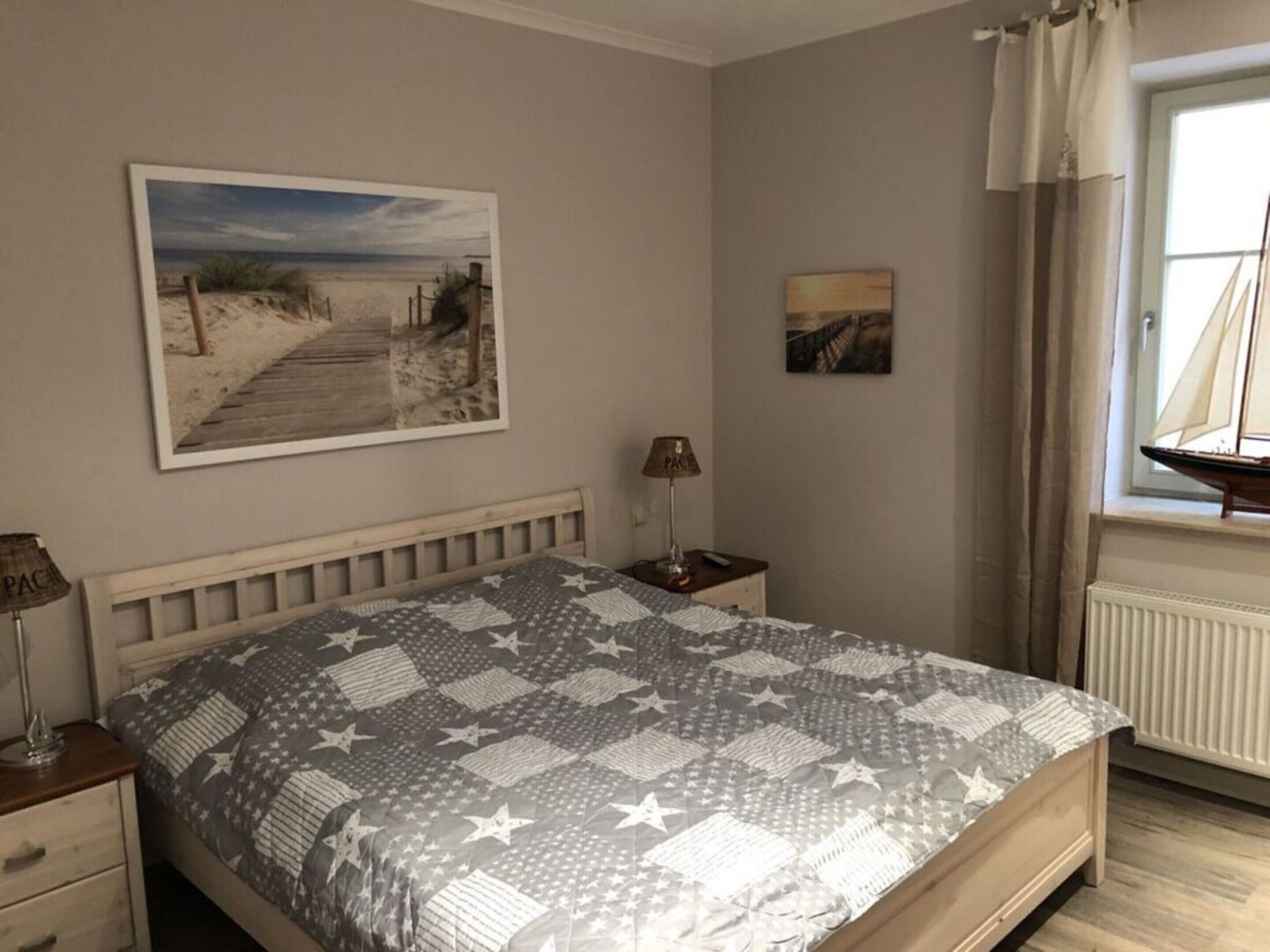 A bedroom with a bed, a nightstand, a lamp, a window with curtains, and two wall-mounted pictures.