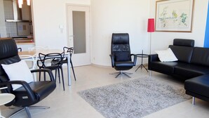 A modern living room with a black leather sofa, a black leather chair, and a dining area with white chairs and a table. - Picturesque Apartment in Nieuwpoort (Nieuwpoort)