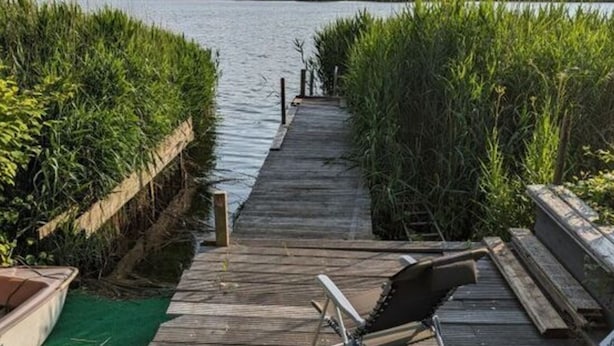 A wooden dock leading to a calm lake, with a rowboat on the left and a lounge chair on the right. - Schleipanorama Modern Retreat (Brodersby)