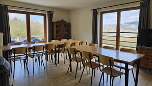 Un espace repas comprenant plusieurs tables et chaises, de grandes fenêtres avec des rideaux, un meuble en bois et un téléviseur.
