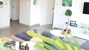 Una sala de estar moderna con sofá, mesa de centro y comedor. Hay cuadros enmarcados en las paredes y un televisor montado en la pared.