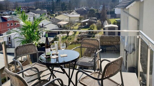 Un balcón con sillas de mimbre y una mesa, con vistas a una zona residencial con casas y una colina al fondo.