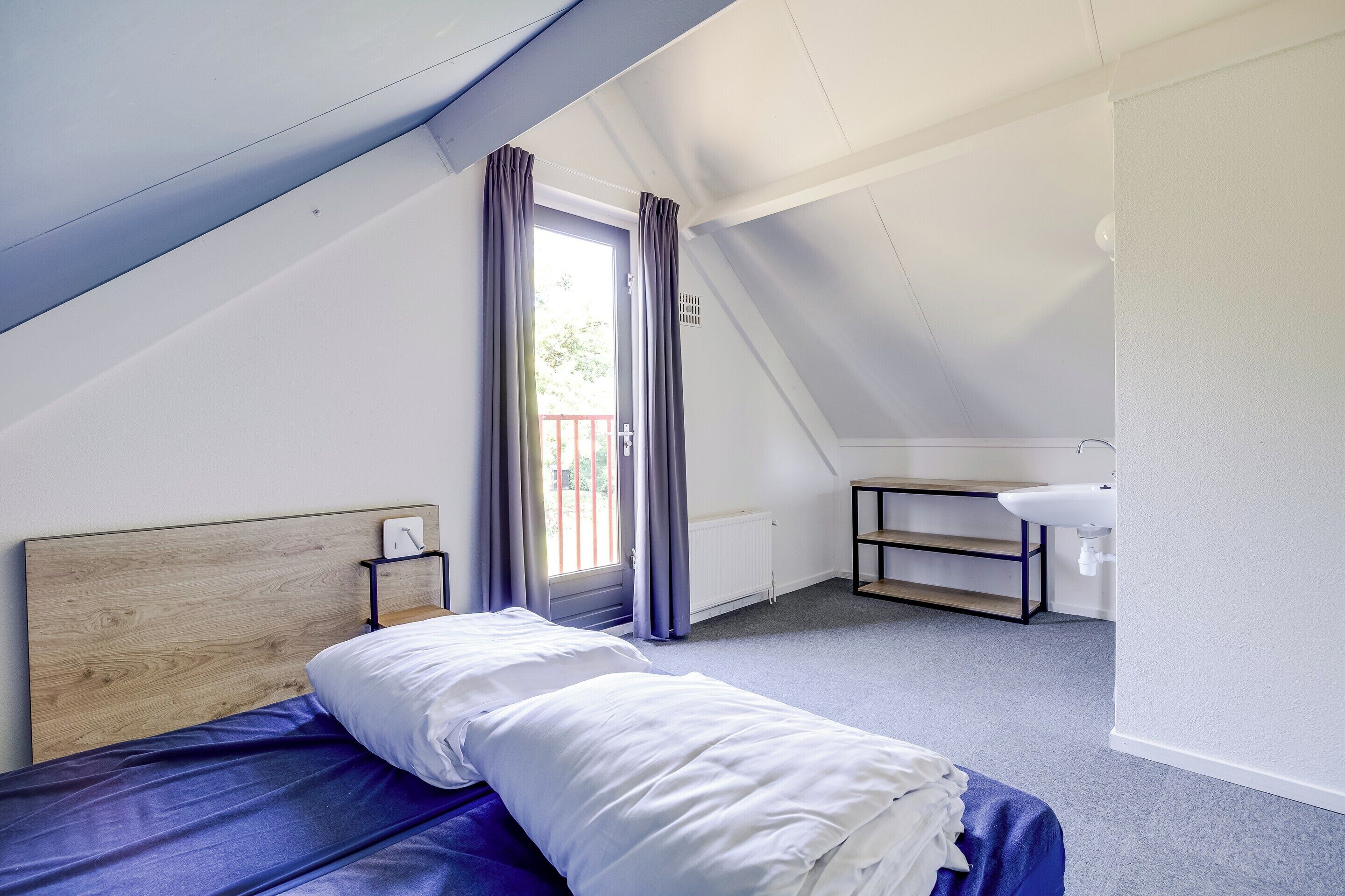 Un dormitorio con techo inclinado, una cama con ropa de cama blanca, una mesita de noche, una ventana con cortinas y un baño con lavamanos y radiador.