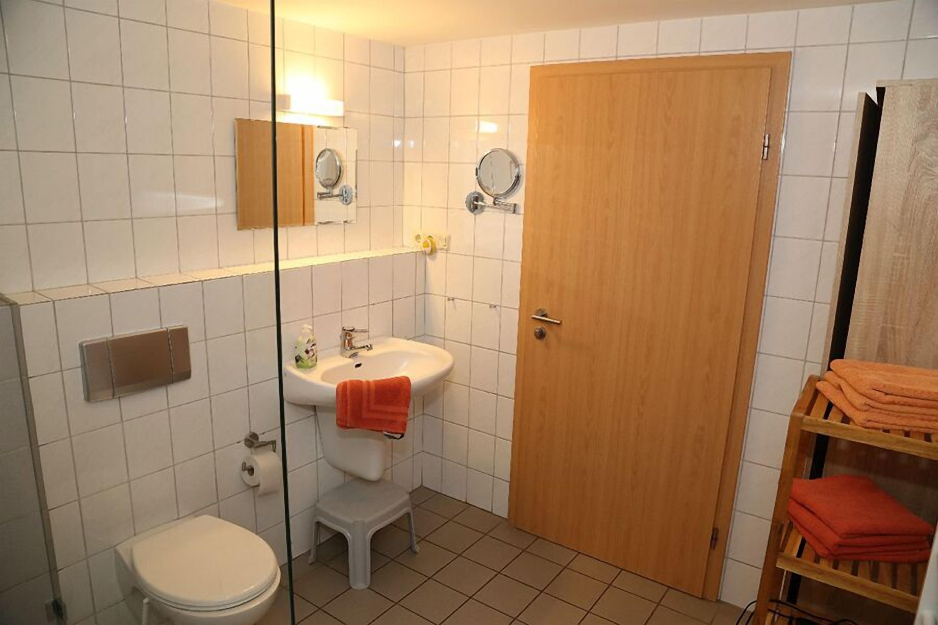 A bathroom with a toilet, sink, and mirror. There is a wooden door and a shelf with folded towels.