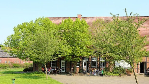 A traditional building with a red-tiled roof, surrounded by lush green trees. - Ferienhof Schwienhorst - Ferienwohnung Bauernstübchen. 1-7 Pers (Warendorf-Hoetmar)