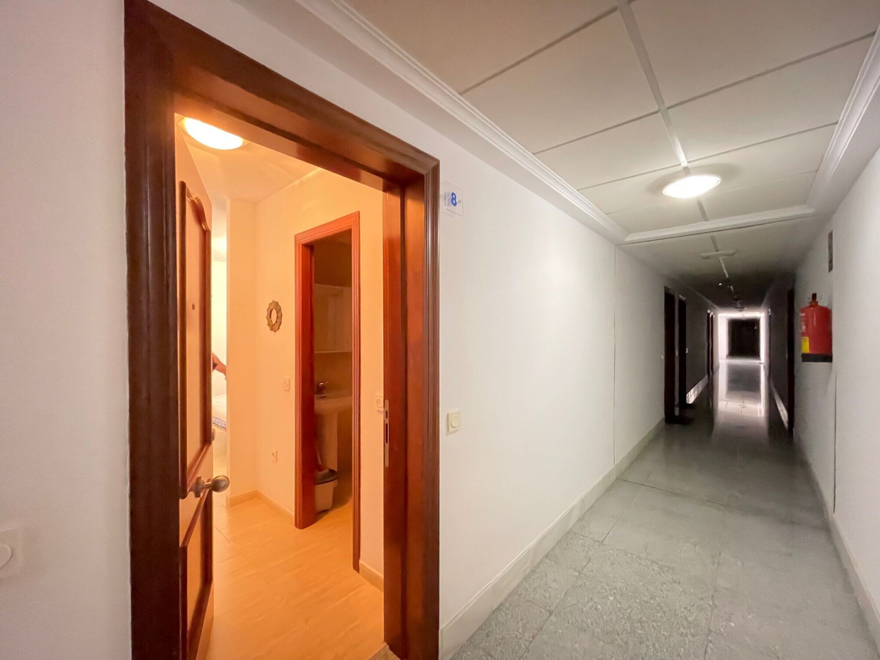 A hotel hallway with a wooden door, a small clock, and a fire extinguisher on the wall.