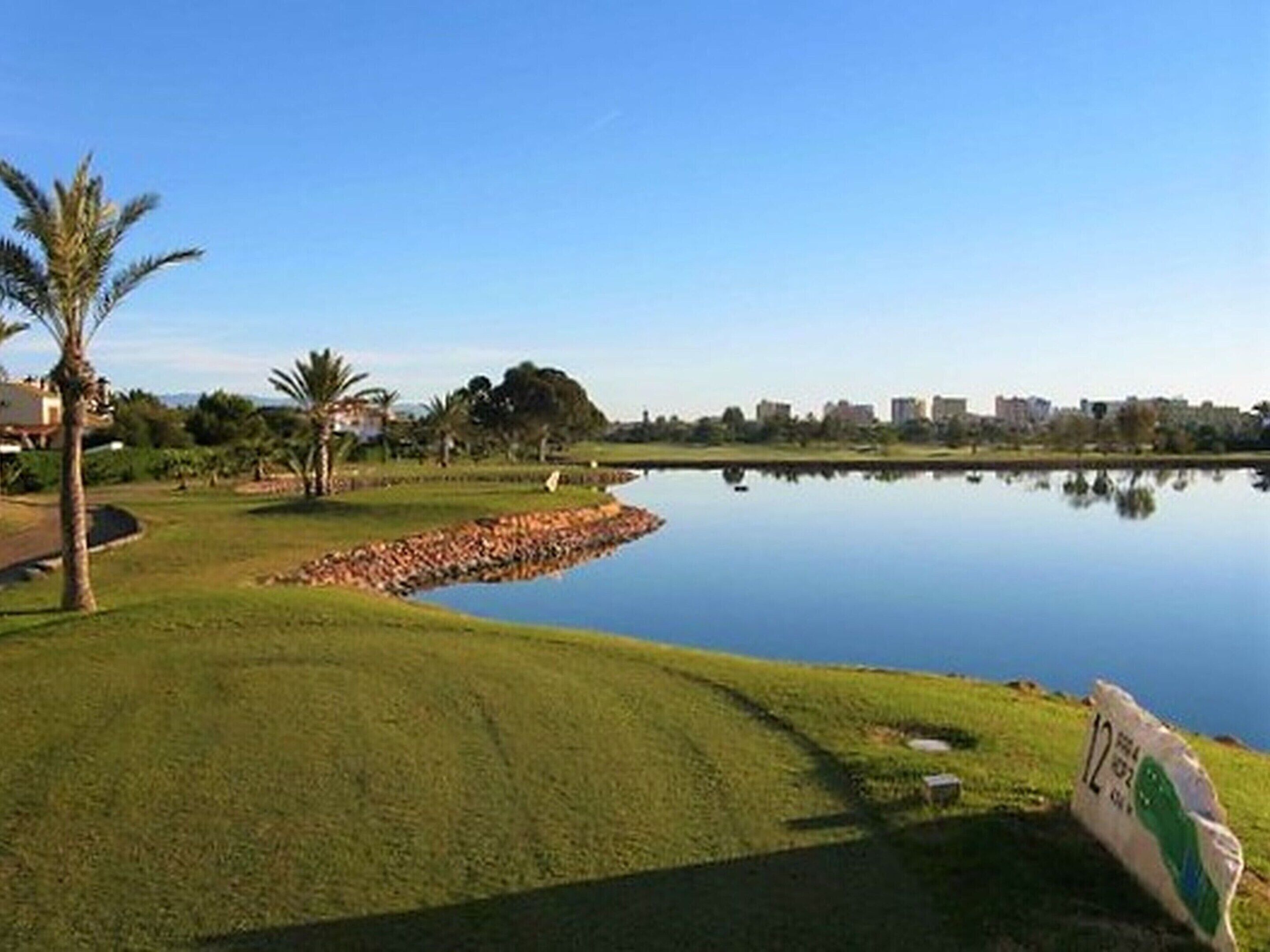 Un campo de golf con un lago tranquilo, un césped cuidado y un horizonte urbano distante.