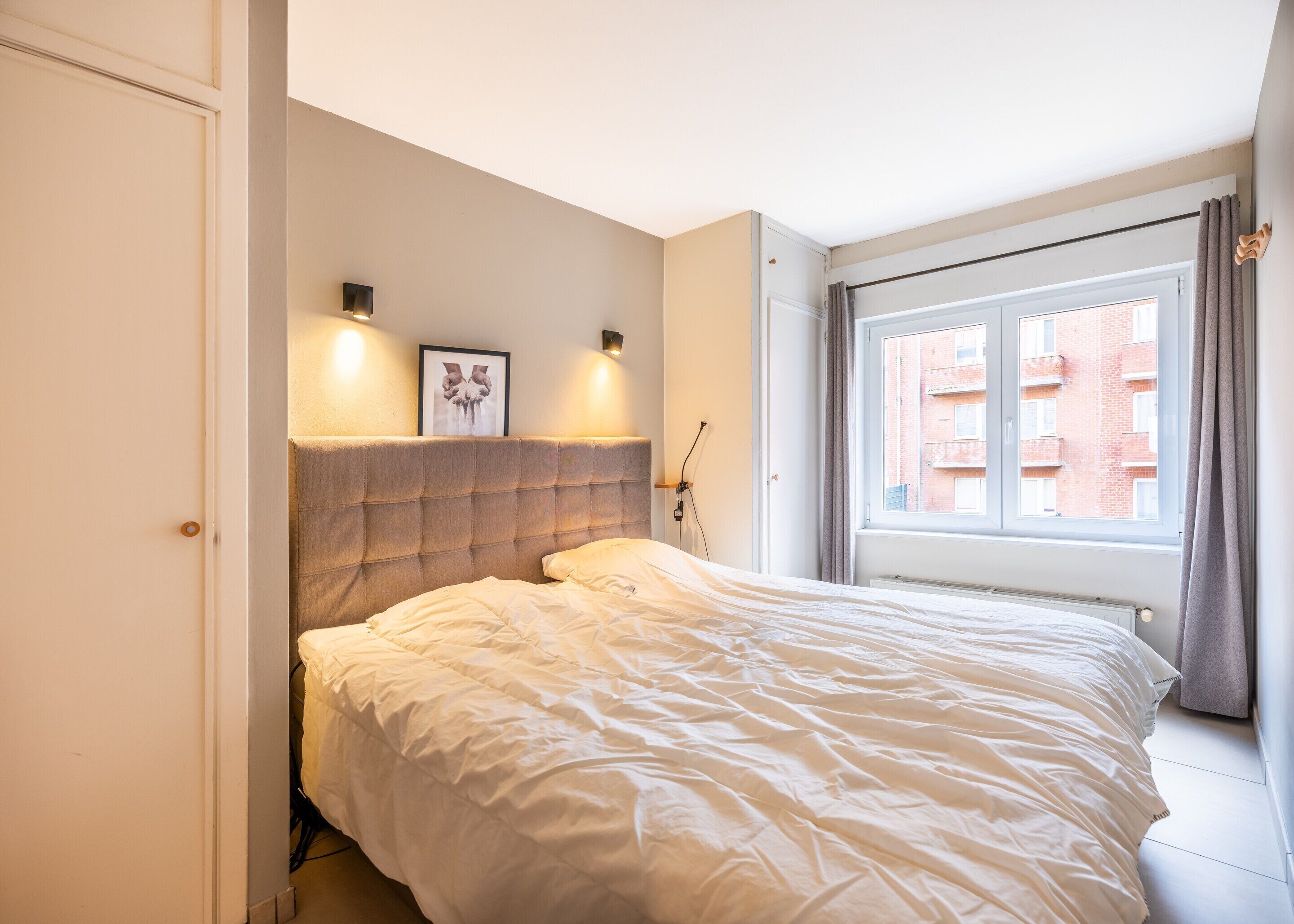 A bedroom with a large bed, a headboard, a framed picture on the wall, and a window with curtains.