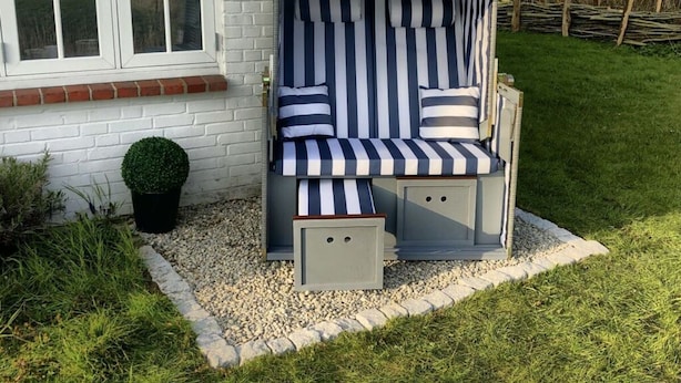 Een terras met strepen, met een bankje en een tafeltje, gelegen op een grindpad naast een witte bakstenen muur.