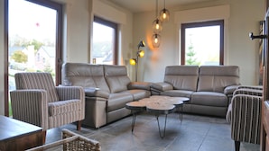A spacious living area with a large L-shaped sectional sofa, two armchairs, and a coffee table. There are large windows providing natural light and a view of the outside. - Villa in Durbuy With Ardennes Views (Durbuy)