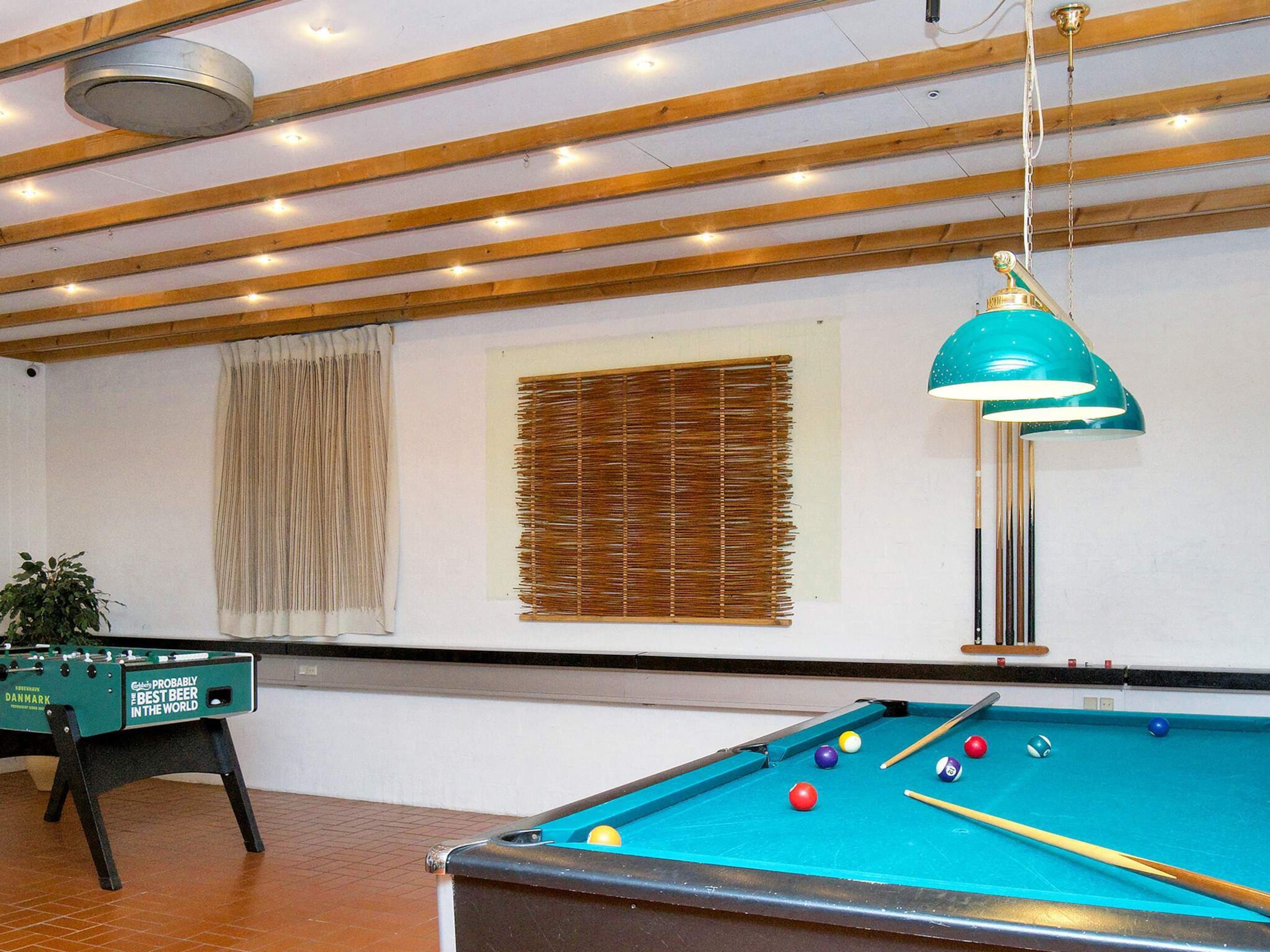 Una mesa de billar con bolas y tacos, una mesa de fútbol de mesa y una lámpara colgante en una habitación con vigas de madera y una ventana con persianas.