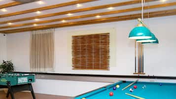Una mesa de billar con bolas y tacos, una mesa de fútbol de mesa y una lámpara colgante en una habitación con vigas de madera y una ventana con persianas.