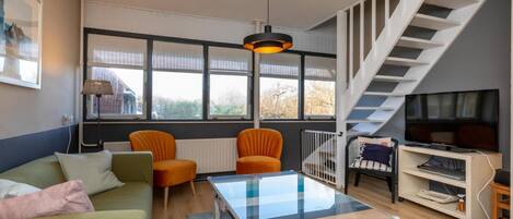 A living room with a sofa, two orange chairs, a glass coffee table, a TV, and a staircase.
