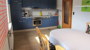 A compact kitchen with blue cabinets, a stove, oven, and a sink. There is a dining table with chairs and a green wall decoration. - Holiday Apartment Heitmann (Hilders)