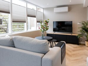 A modern living room with a grey sofa, a TV on a black stand, a small round table, and a potted plant. - Holiday Apartment Costa (Callantsoog)