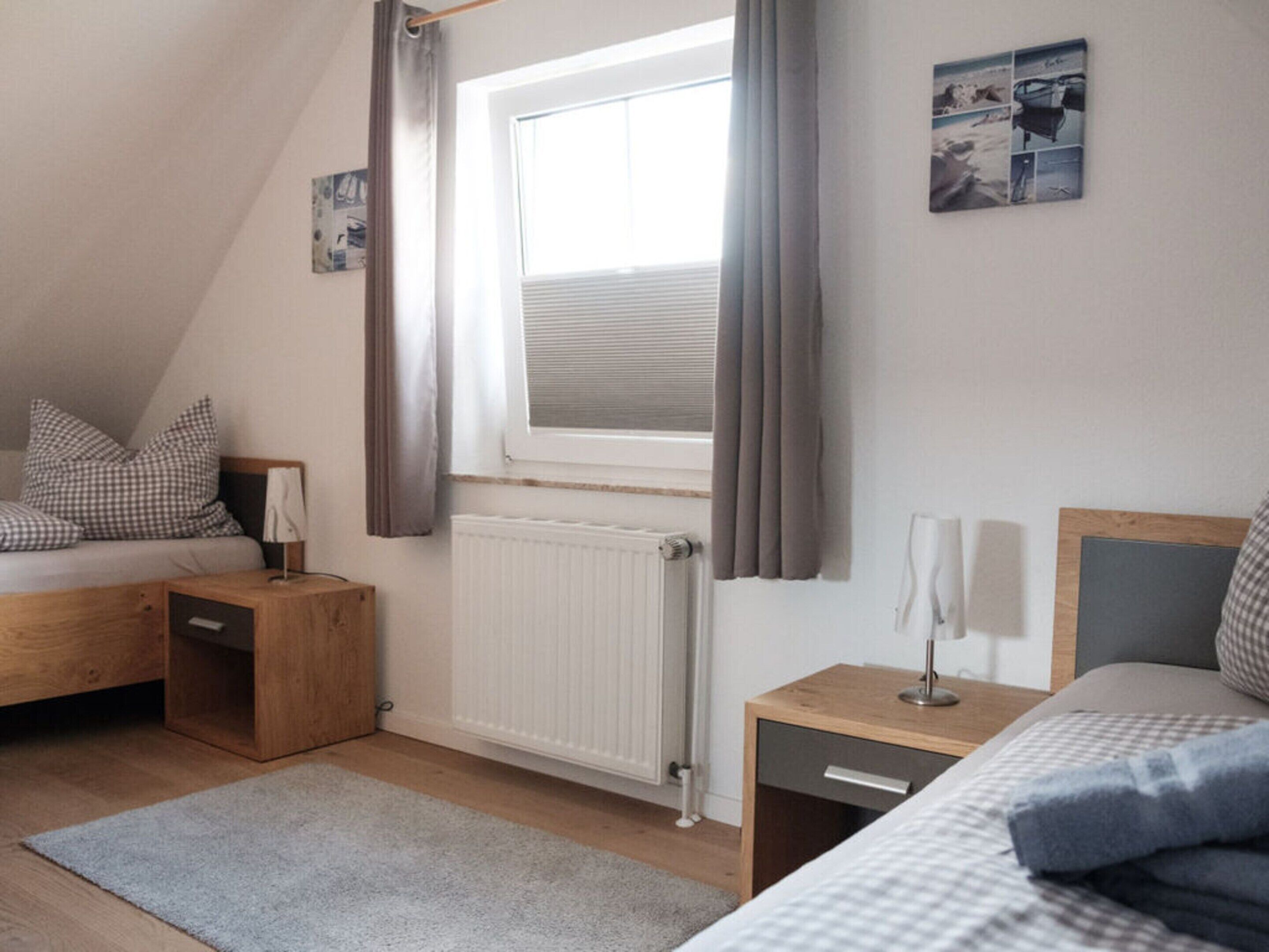 Ein Schlafzimmer mit schrägen Wänden, einem Fenster mit Jalousien, einem Heizkörper, einem Nachttisch mit Lampe und einem Bett mit karierter Bettwäsche.