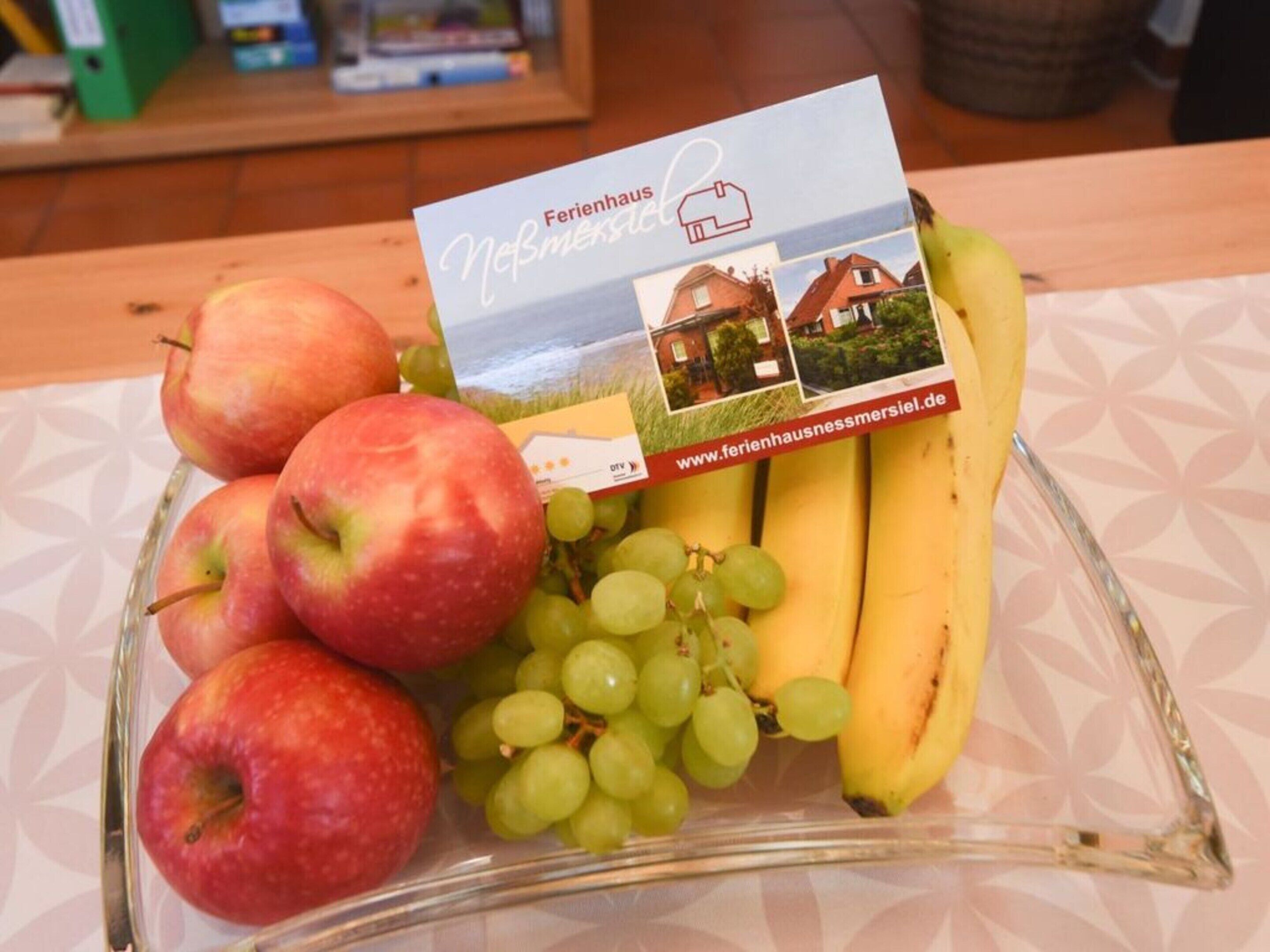 A fruit platter with apples, grapes, and bananas, and a business card for Ferienhaus Nebmenschiel.