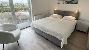 A modern bedroom with a large bed, a chair, and a view through a sliding glass door. - Holiday Home in Zeeland With Workation Setup (Wemeldinge)
