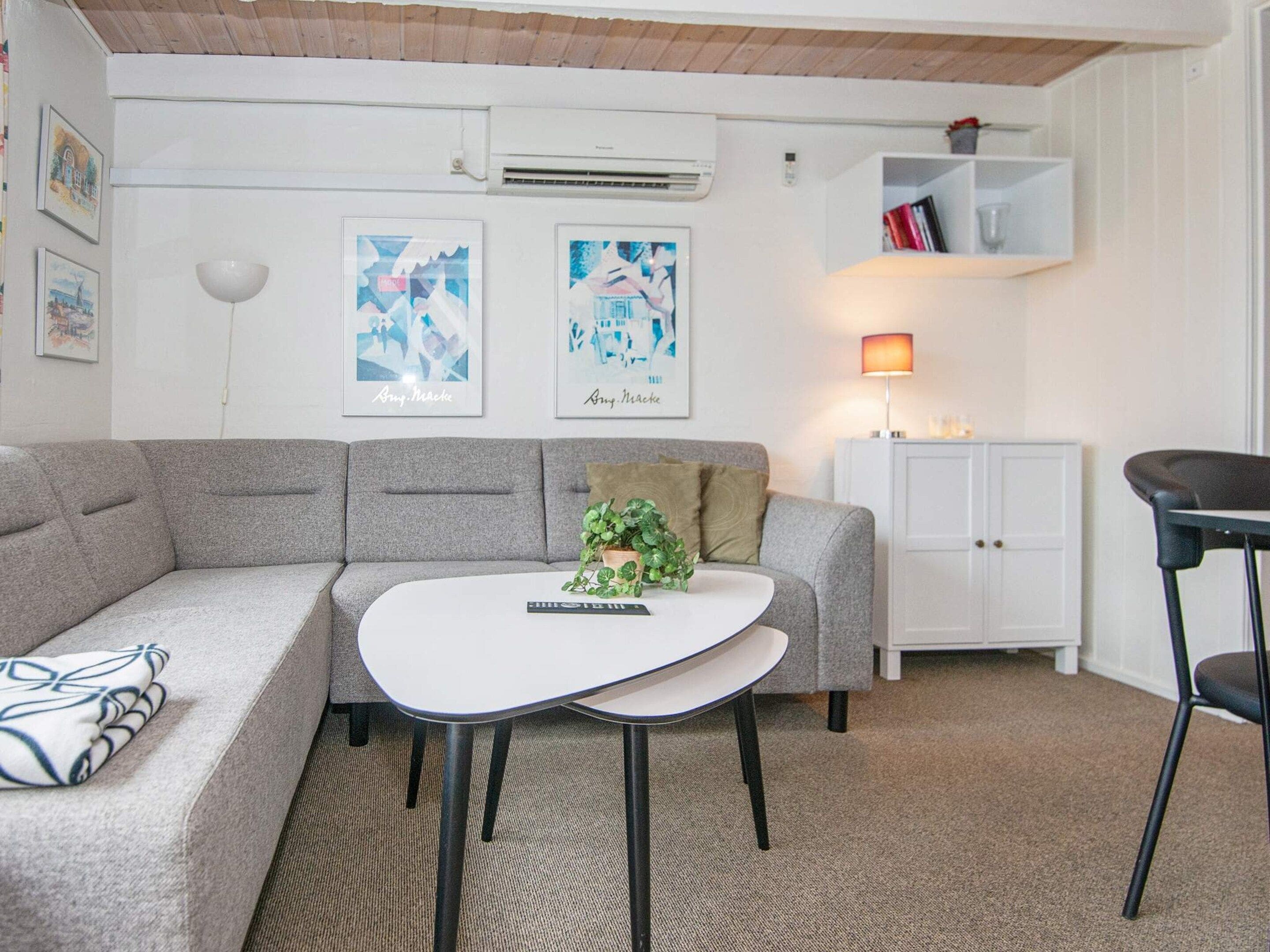 Una sala de estar moderna con un sofá gris, una mesa de centro blanca y una silla negra. Hay un armario blanco y una estantería con libros y un vaso.