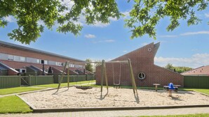 A playground with swings and a slide, surrounded by a brick building and a green fence. - 4 Person Holiday Park Home in Hemmet (Hemmet)