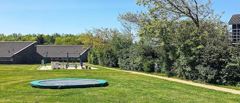 A grassy outdoor area with a trampoline, a building with a sloped roof, and a pathway leading to it.