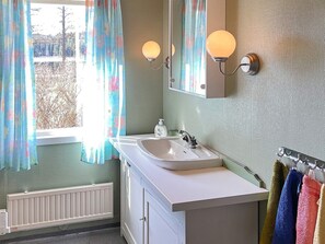 A bathroom with a white sink, a mirror, and two wall-mounted lights. There is a window with floral curtains, and towels are hanging on a rack. - 4 Star Holiday Home in Askersund (Askersund)