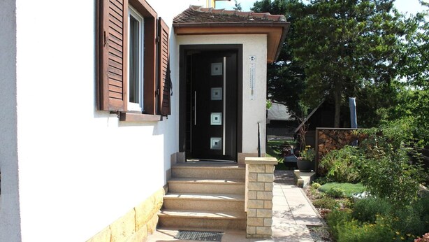 A building with a white exterior, a brown door, and a set of stairs leading to the entrance. - Ferienwohnung in Lichtenhain (Sebnitz)