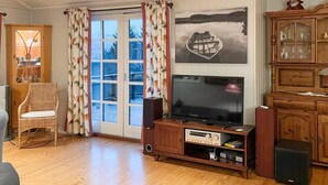Een woonkamer met een houten vloer, een televisie op een houten dressoir, een rieten stoel, een kast met glazen deuren en een raam met gordijnen.