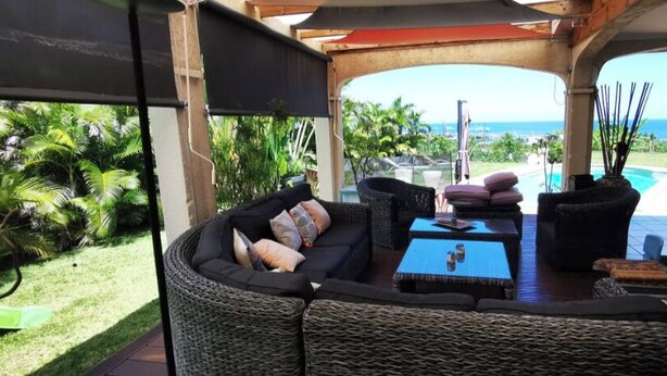 A covered outdoor seating area with wicker furniture, a pool, and a view of the ocean. - Location Villa Juin-juillet (La Possession)