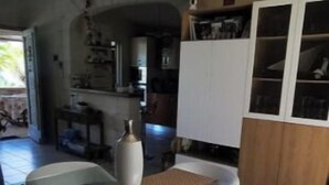 A modern dining area with a glass table and white chairs, a kitchen in the background, and a tiled floor. - Location Villa Juin-juillet (La Possession)