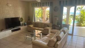 A living room with a TV, sofa, coffee table, and a view of the outdoors. - Location Villa Juin-juillet (La Possession)