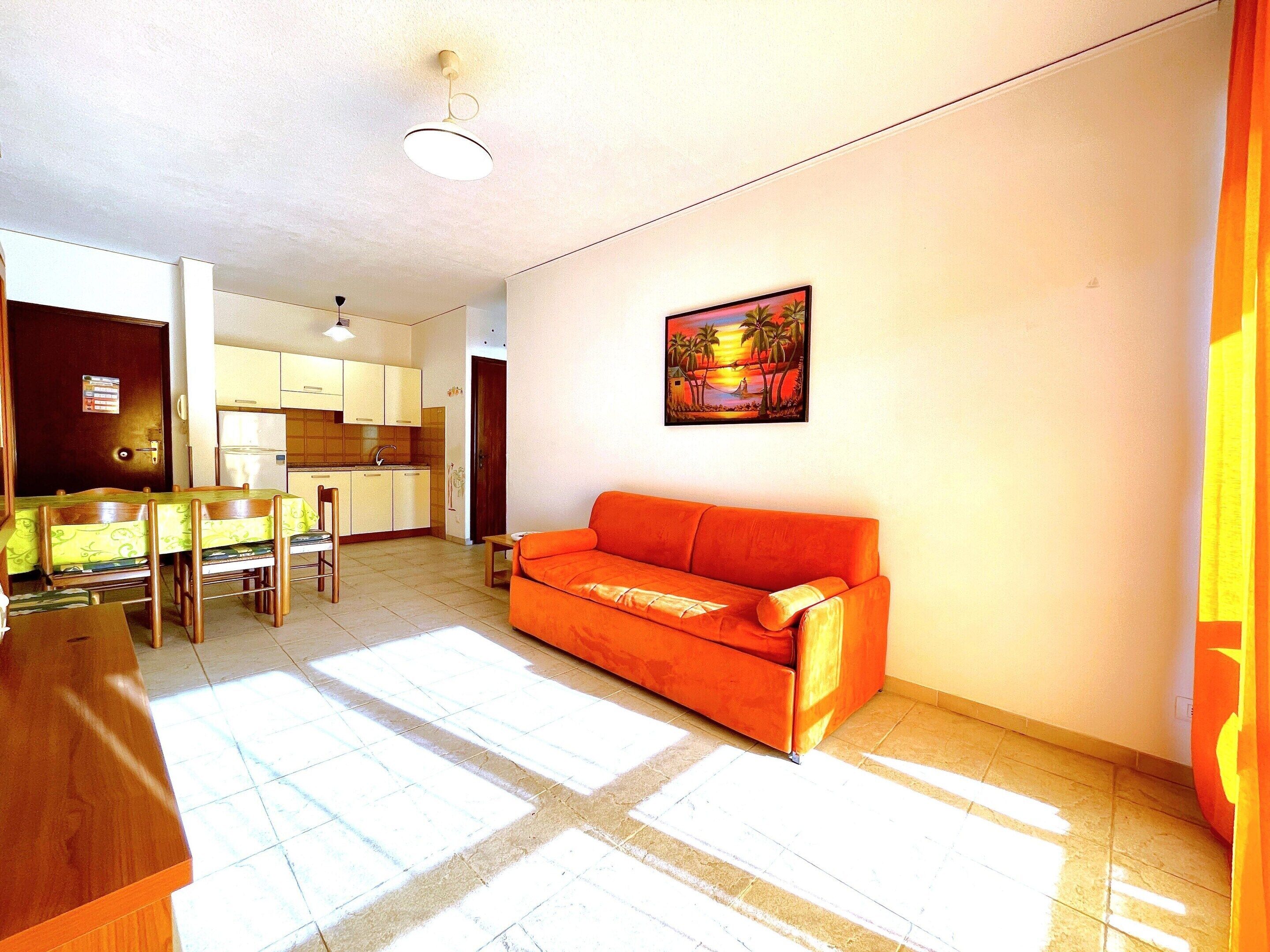 A living room with a sofa, a dining table with chairs, a kitchen area, and a painting on the wall.