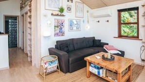 A cozy living room with a dark grey sofa, a wooden coffee table, and a bookshelf. There are framed artworks on the walls, a window with a view of greenery, and a door leading to another room. - 5 Star Holiday Home in Glesborg (Glesborg)