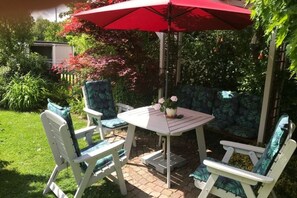 Un patio con mesa y sillas, rodeado de abundante follaje y un paraguas rojo.