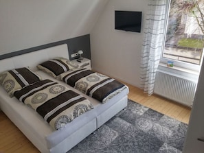 Ein Schlafzimmer mit Bett, Nachttisch, einem an der Wand montierten Fernseher und einem Fenster mit Vorhängen.