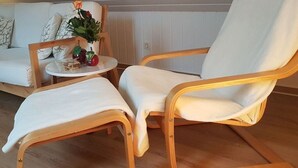 Habitació amb un sofà blanc, dues cadires de fusta, una taula rodona petita amb un ram de flors i un radiador.