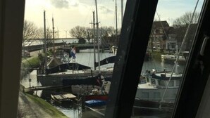 Une vue à travers une fenêtre sur une marina où sont amarrés des bateaux, avec un ciel dégagé et des bâtiments en arrière-plan.